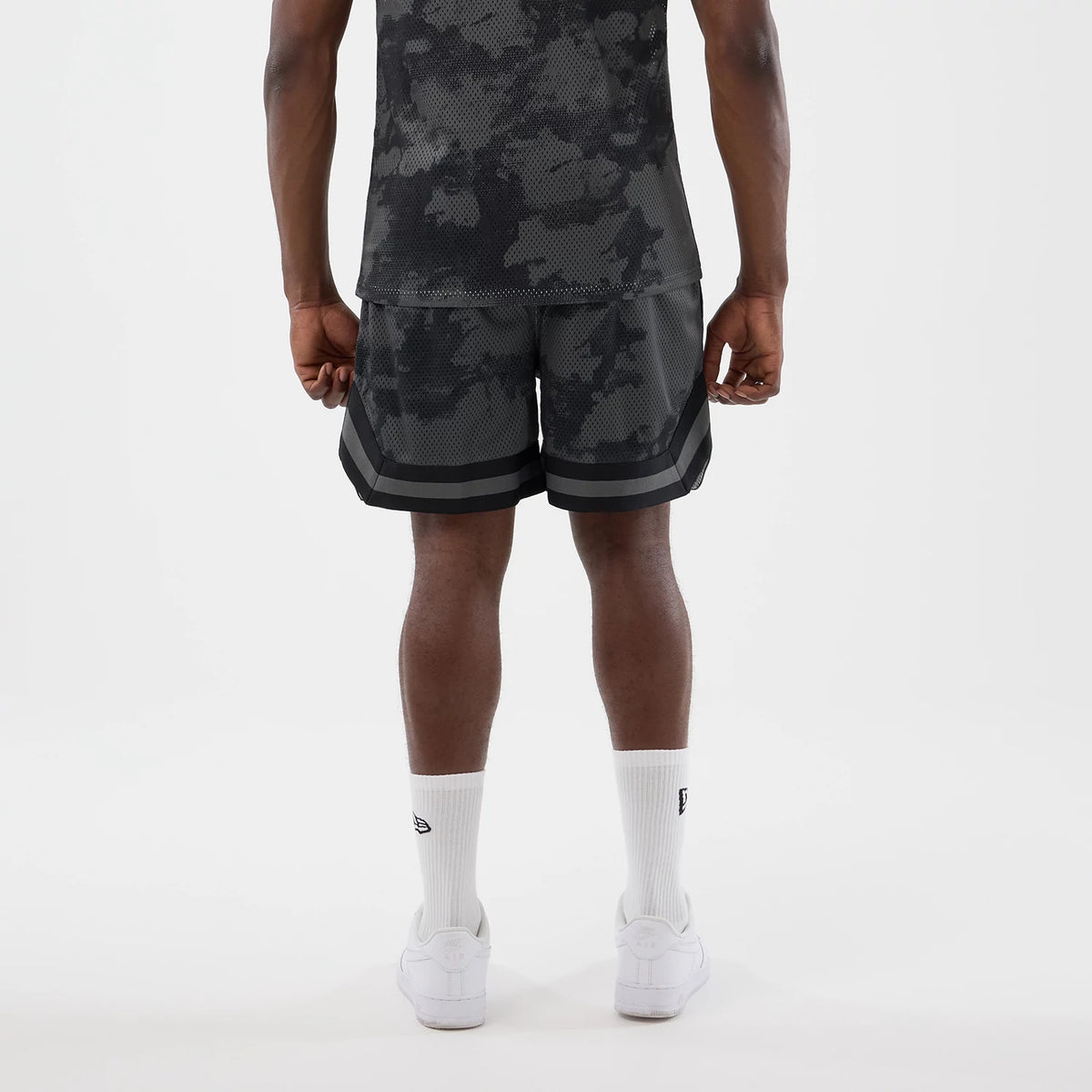New Era All Over Print Mesh Black Camo Shorts, back view on a model showing a camo pattern with two horizontal black stripes near the hem, paired with a matching camo top, highlighting the relaxed fit and structured shorts design.