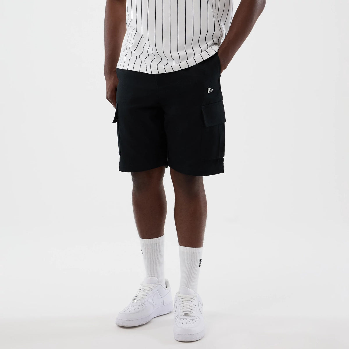 New Era Black Cargo Shorts, front view on a model styled with a pinstriped shirt and white trainers, displaying spacious cargo pockets on each side and a distinctive embroidered New Era logo on the left front pocket.