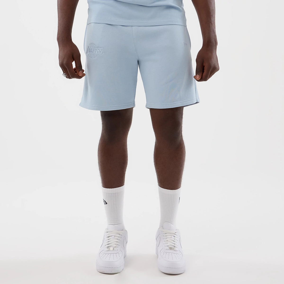 New Era LA Lakers NBA League Essential Pastel Blue Shorts, front view on-model showing an embroidered Lakers logo on the left thigh, pastel blue colour throughout, and styled with a tucked-in top, white socks, and white trainers for a casual look.