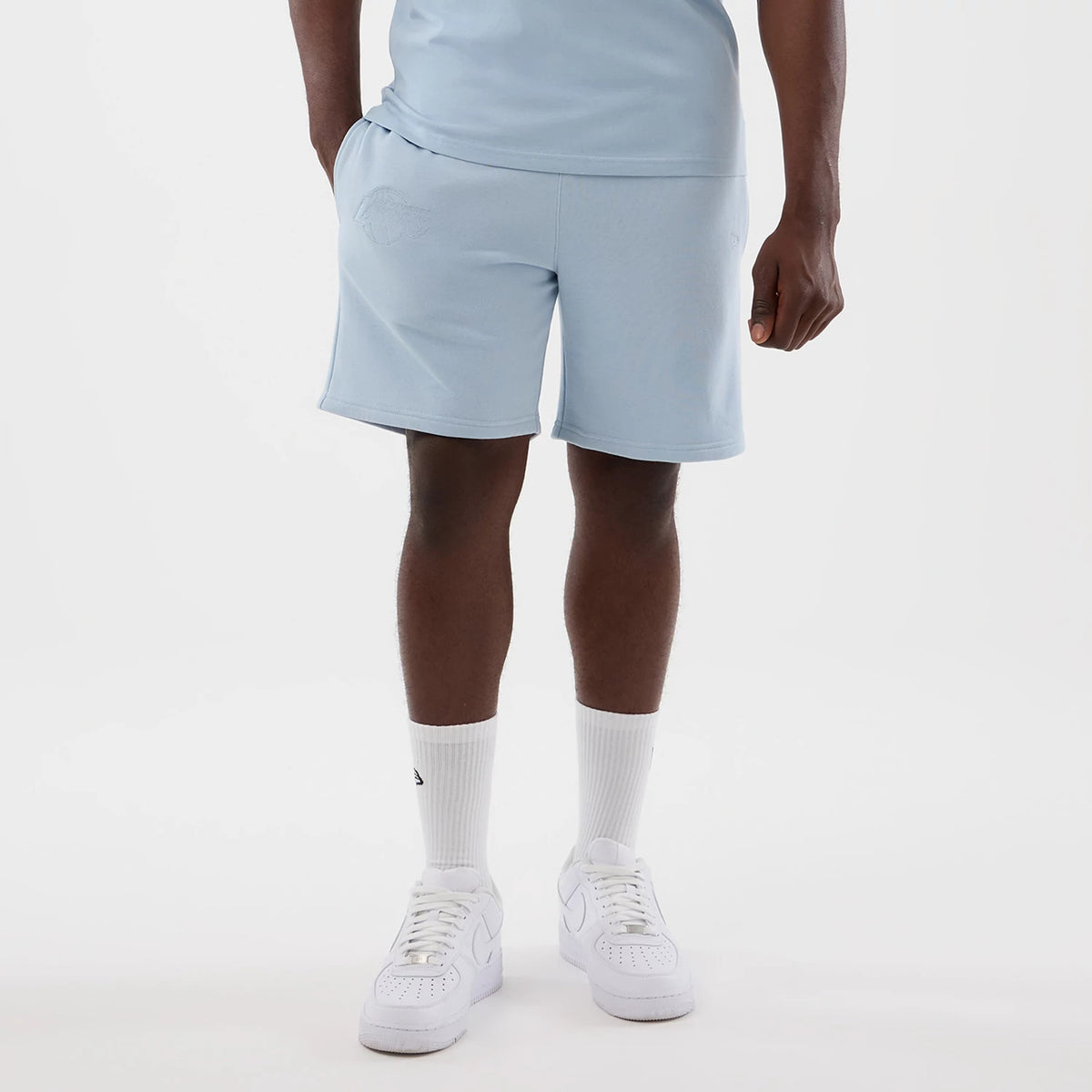 New Era LA Lakers NBA League Essential Pastel Blue Shorts, front view on a model showcasing the pastel blue shorts with an embroidered LA Lakers logo on the left leg, styled with a matching pastel blue top and white trainers.