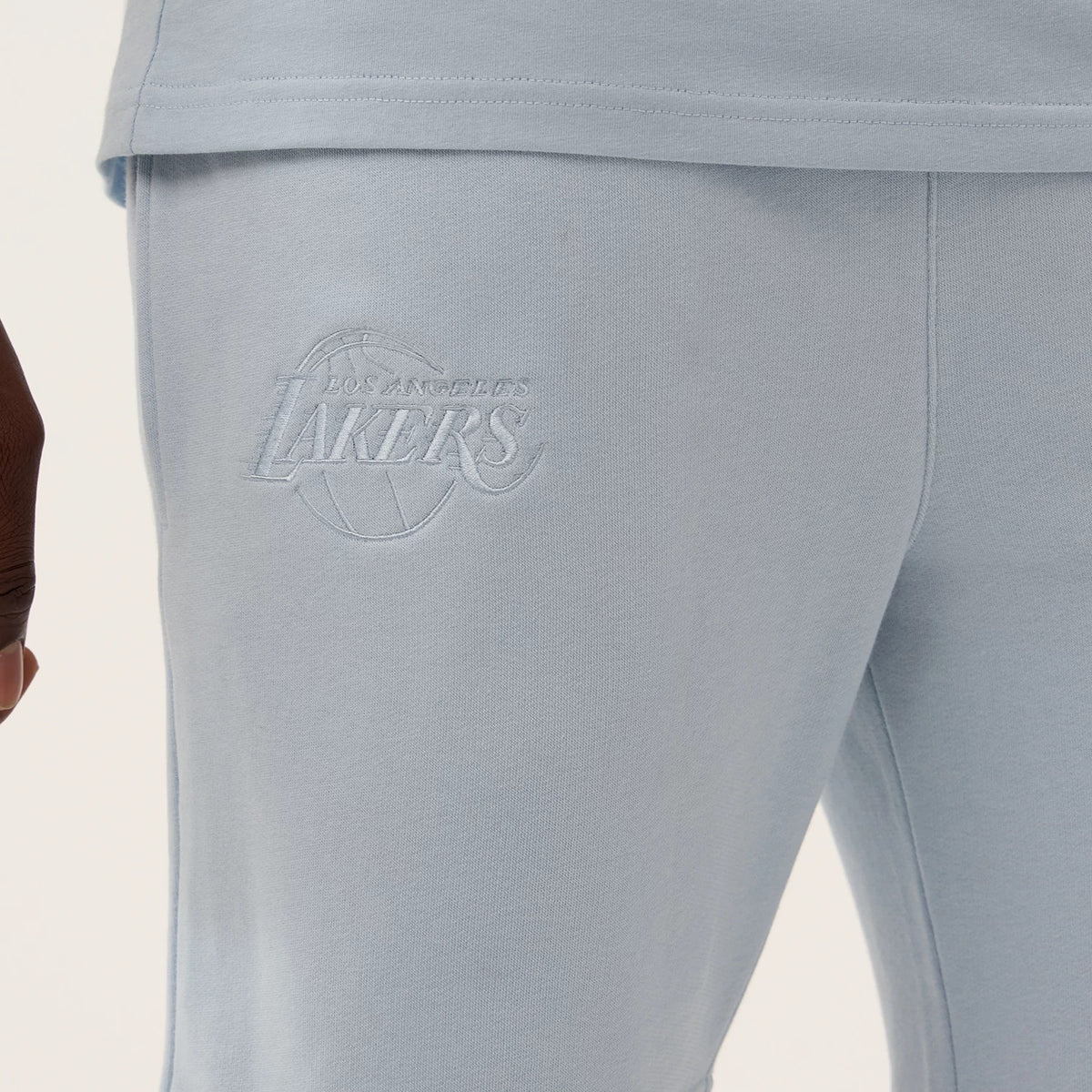 New Era LA Lakers NBA League Essential Pastel Blue Shorts, side view close-up showing the finely embroidered Los Angeles Lakers logo on the pastel blue fabric above the knee, paired with a matching top at the hem.