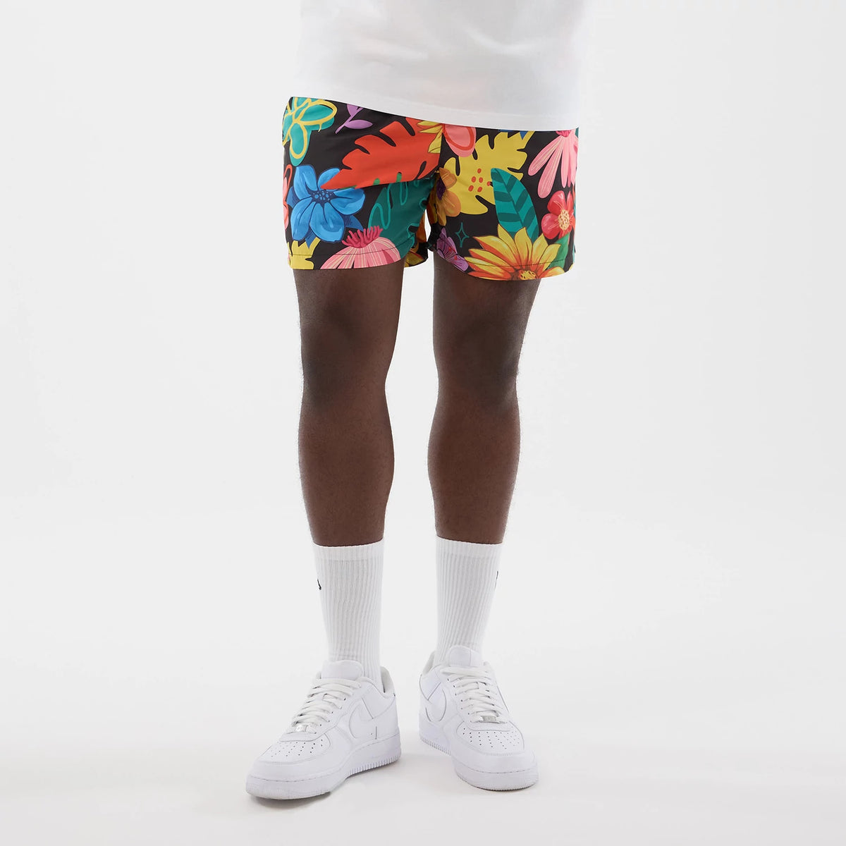 New Era Floral Black Swimming Shorts, front view on a model showing a floral pattern in blue, pink, and yellow against a black background, with an above-the-knee length and styled with white socks and trainers.
