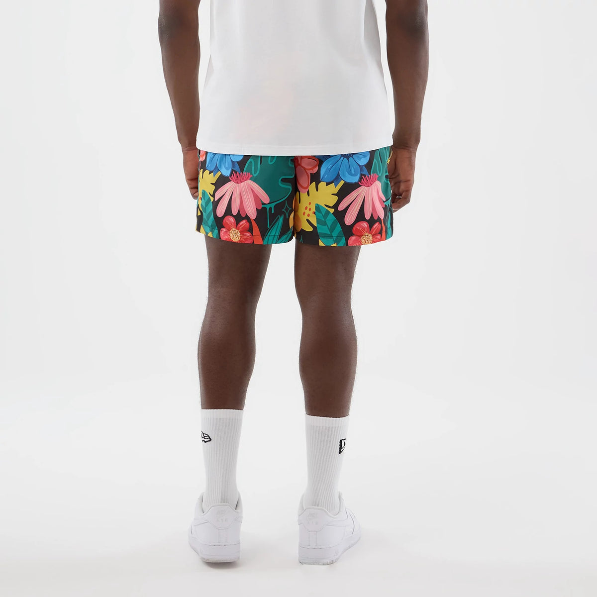 New Era Floral Black Swimming Shorts, back view on a model featuring a floral print with large pink, blue and red flowers on a black background, complemented by a plain white T-shirt, white socks, and white trainers.