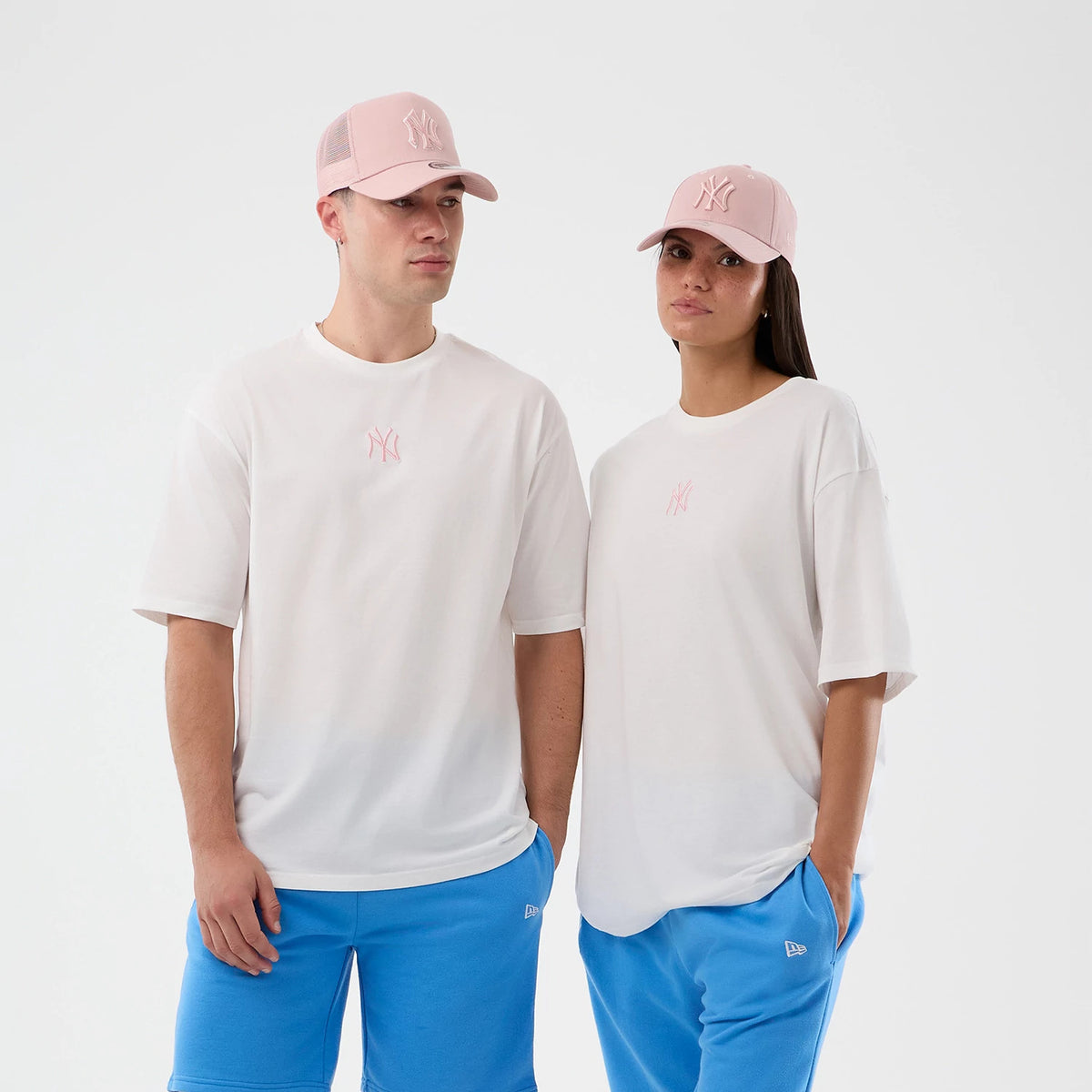 New Era New York Yankees MLB Midi League Essential White Oversized T-Shirt, on-model view highlighting a relaxed fit with the light pink embroidered Yankees logo centred on the chest, paired with bright blue trousers and matching light pink caps.