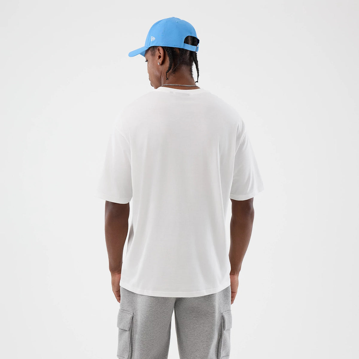 New Era Athletics MLB League Essential White Oversized T-Shirt, back view on a model with a light blue cap, showing the plain white back of the oversized T-shirt and short sleeves, styled with light grey cargo shorts for a casual look.