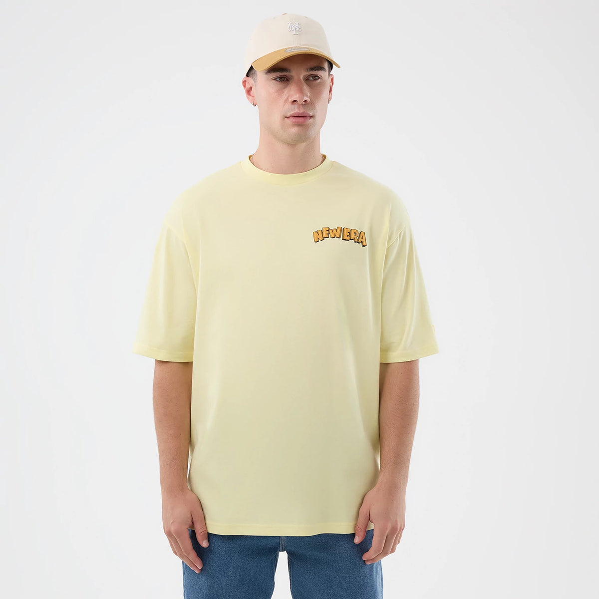 New Era Fun Beach Party Graphic Pastel Yellow Oversized T-Shirt, front view on-model showing a pastel yellow design with a orange embroidered New Era logo on the left chest, and relaxed short sleeves over blue jeans.