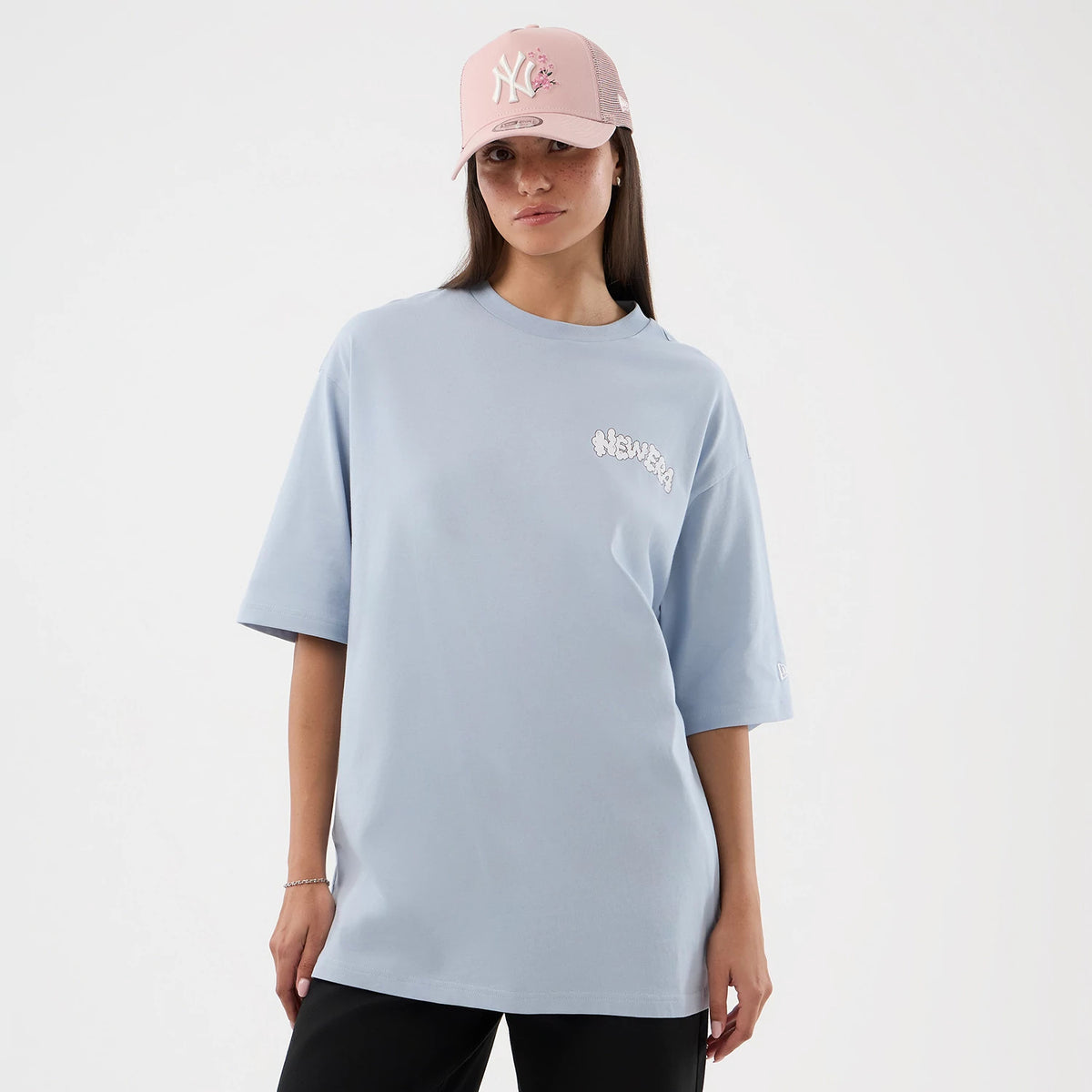 New Era Fun Block Party Graphic Pastel Blue Oversized T-Shirt, front view on-model showing the New Era logo in white on the chest, loose short sleeves with a small embroidered logo at the edge, paired with a pink cap featuring a white NY logo.