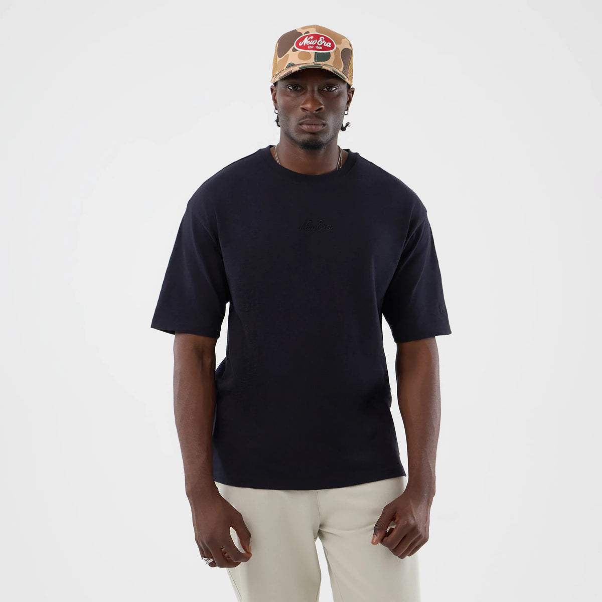 New Era Slub Navy Oversized T-Shirt, front view on a model showcasing the classic navy tone, featuring a embroidered New Era logo on the chest, loose fit short sleeves, and styled with light coloured trousers and matching camo patterned cap.