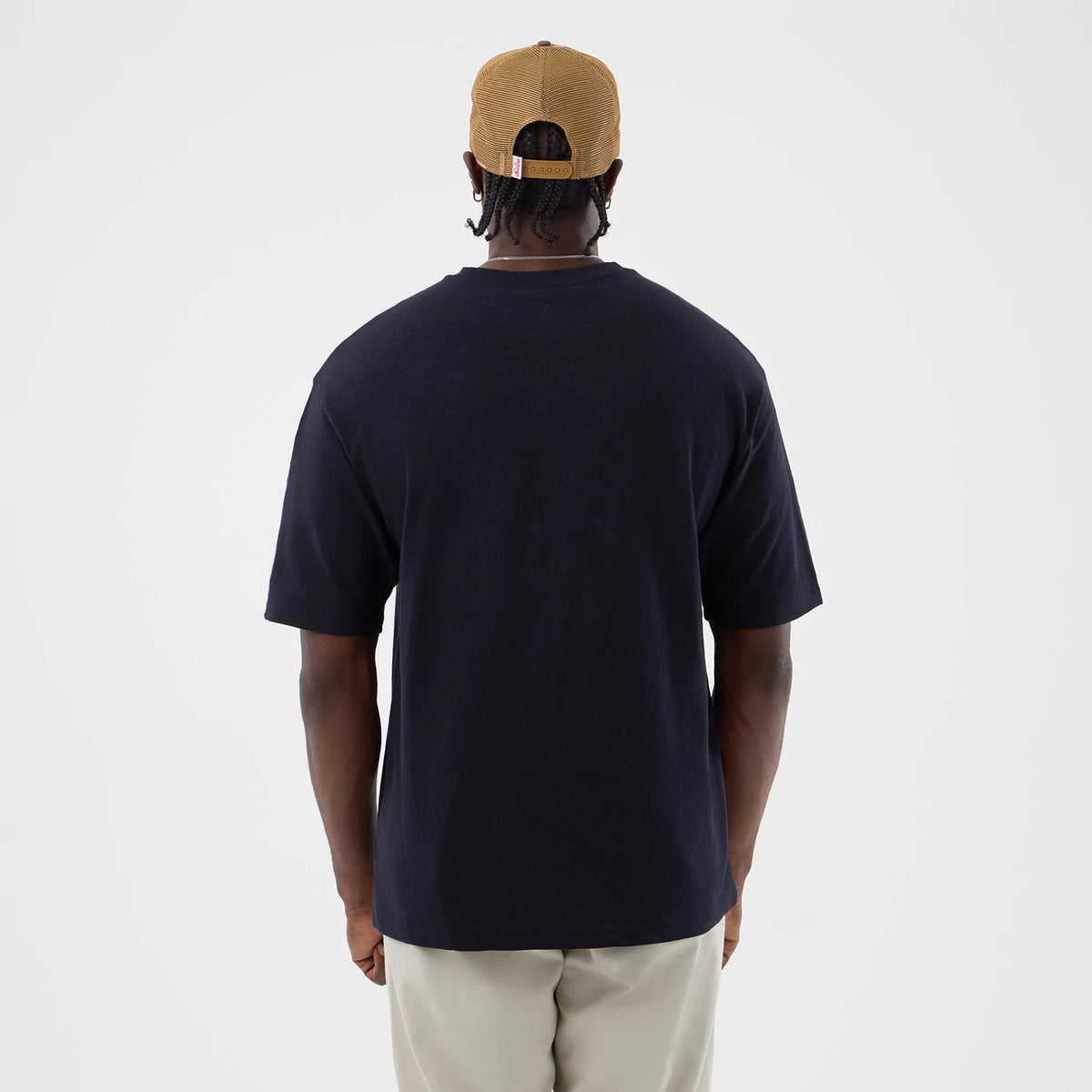 New Era Slub Navy Oversized T-Shirt, back view on a model wearing beige trousers and a tan cap, showing the plain navy back with short sleeves ending at the elbow, providing a clean and simple aesthetic.