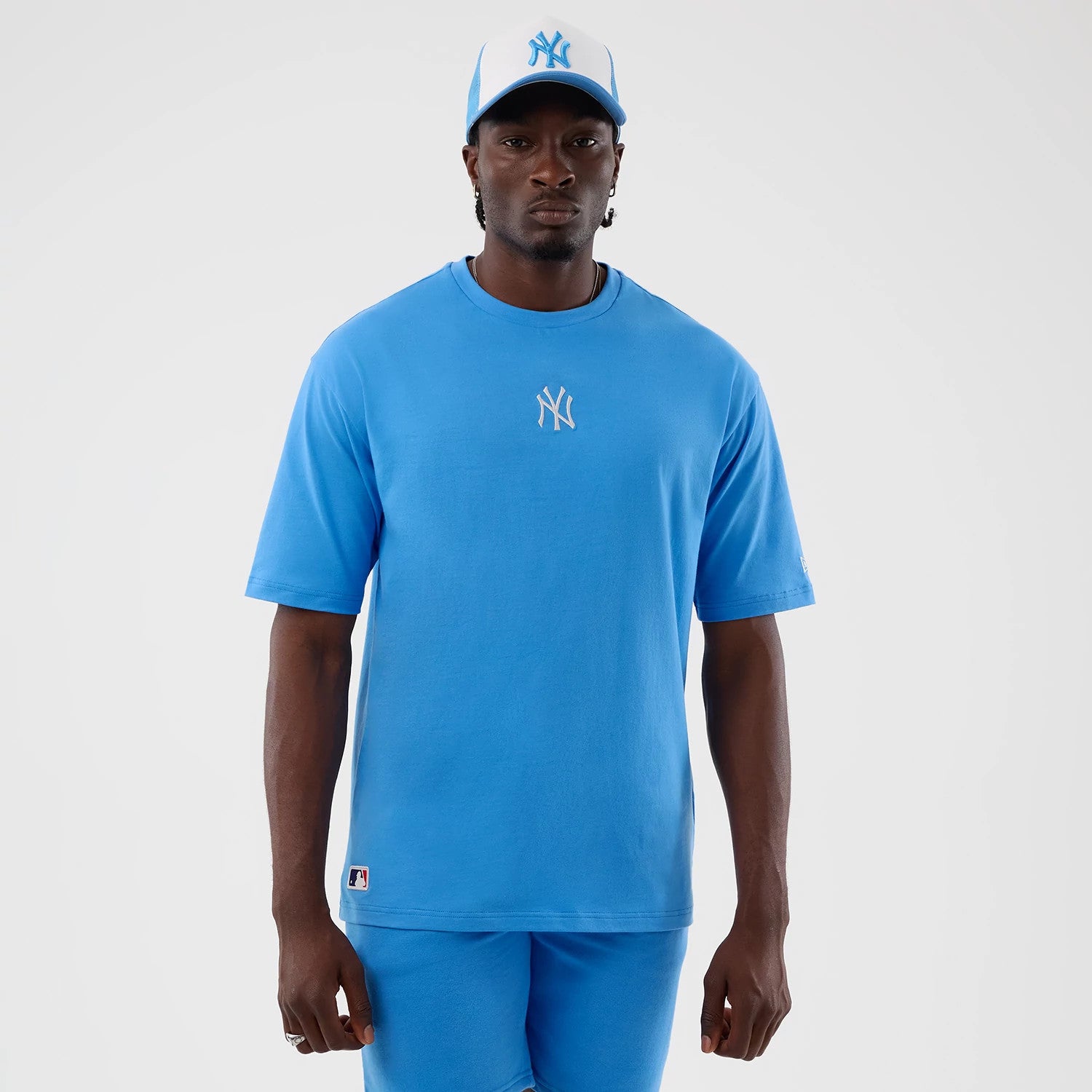 New Era New York Yankees MLB League Essential Pastel Blue Oversized T-Shirt, front view on a model styled with a matching cap, featuring a white embroidered Yankees logo on the chest and a small MLB logo near the hem on the left side.