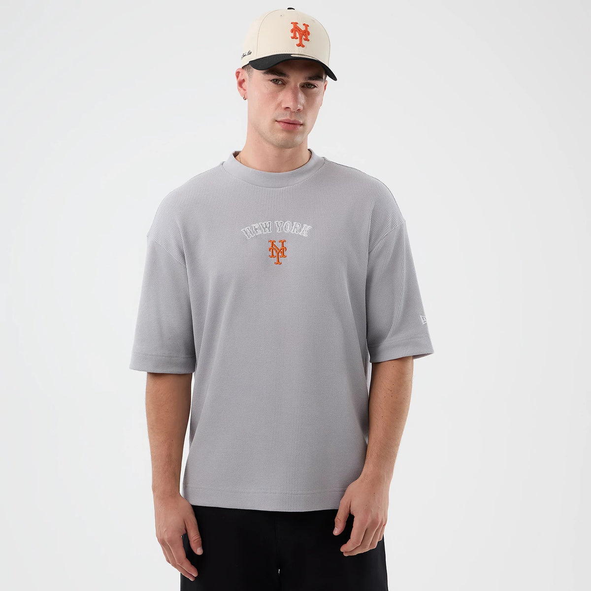 New Era New York Mets MLB Waffle Grey T-Shirt, front view on a model showing the white embroidered New York text and orange Mets logo on the chest, with a New Era logo on the left sleeve, paired with black trousers and a cream cap.