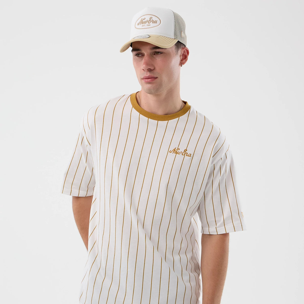 New Era Pinstripe White Oversized T-Shirt, front view on-model featuring thin mustard pinstripes running vertically, a mustard embroidered New Era logo on the left chest, and a contrasting mustard neckline paired with a mesh cap.