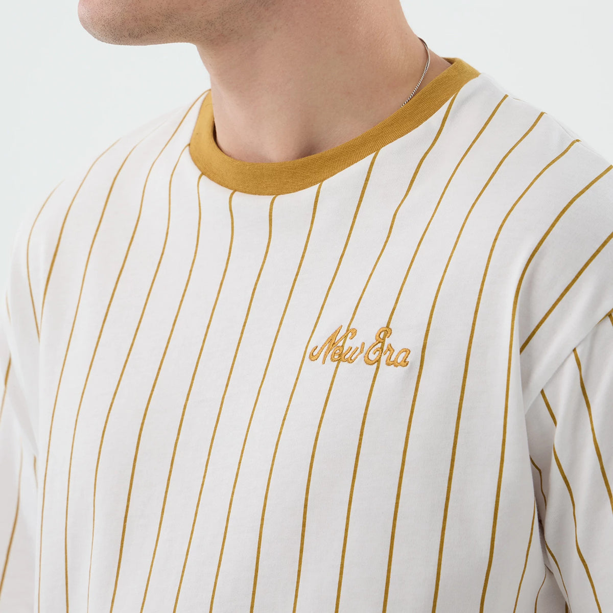 New Era Pinstripe White Oversized T-Shirt, front view showing a white base with golden pinstripes, a contrasting golden collar, and an embroidered New Era logo on the upper left chest, angled across the model's shoulder.