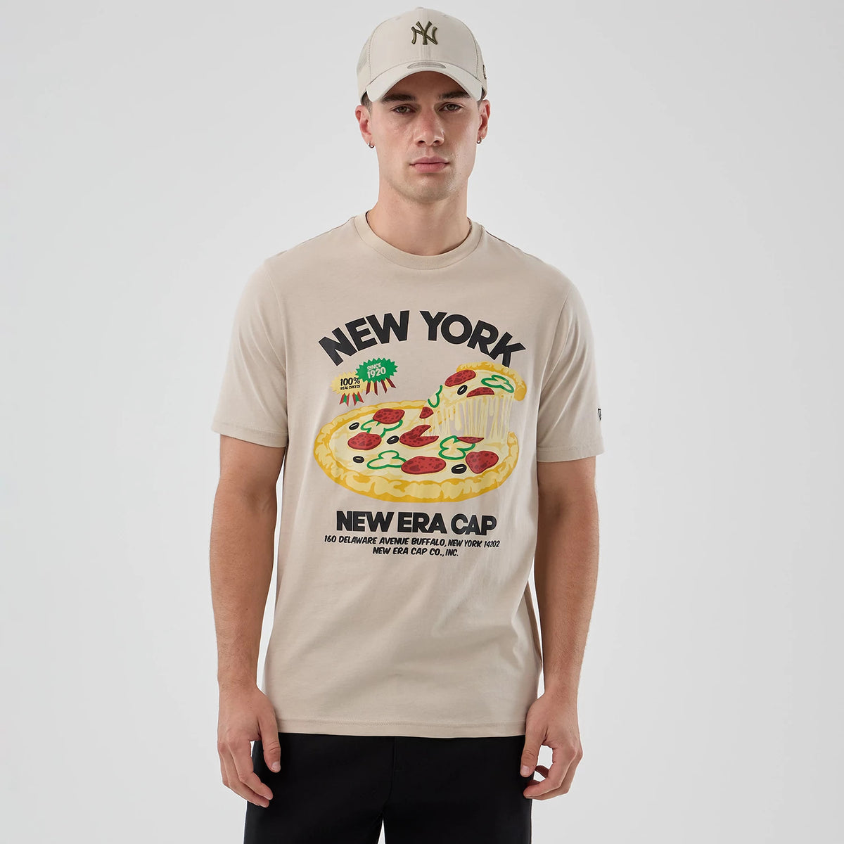 New Era Pizza Graphic Light Beige T-Shirt, front view on-model showing a large colourful pizza graphic with New York in black lettering above, New Era Cap text below, and a small embroidered New Era logo on the left sleeve.