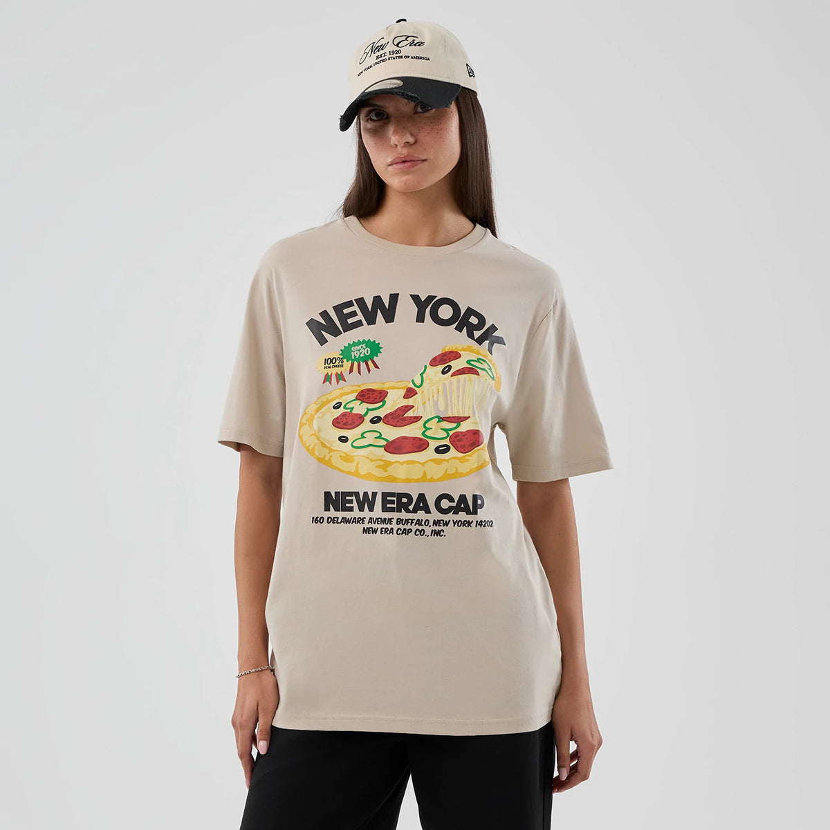 New Era Pizza Graphic Light Beige T-Shirt, front view showing a large pizza graphic with pepperoni and vegetables beneath New York lettering, worn by a model also styled with a beige cap featuring an embroidered New Era logo.