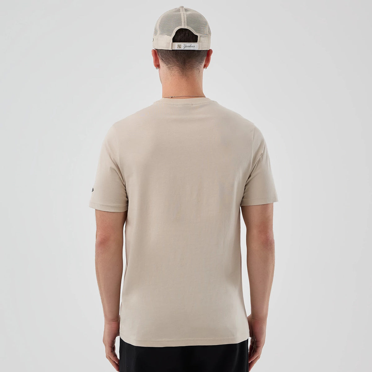New Era Pizza Graphic Light Beige T-Shirt, back view on a model with a mesh cap showing the plain light beige back, short sleeves with a small embroidered New Era logo on the left sleeve near the hem, styled with black trousers.