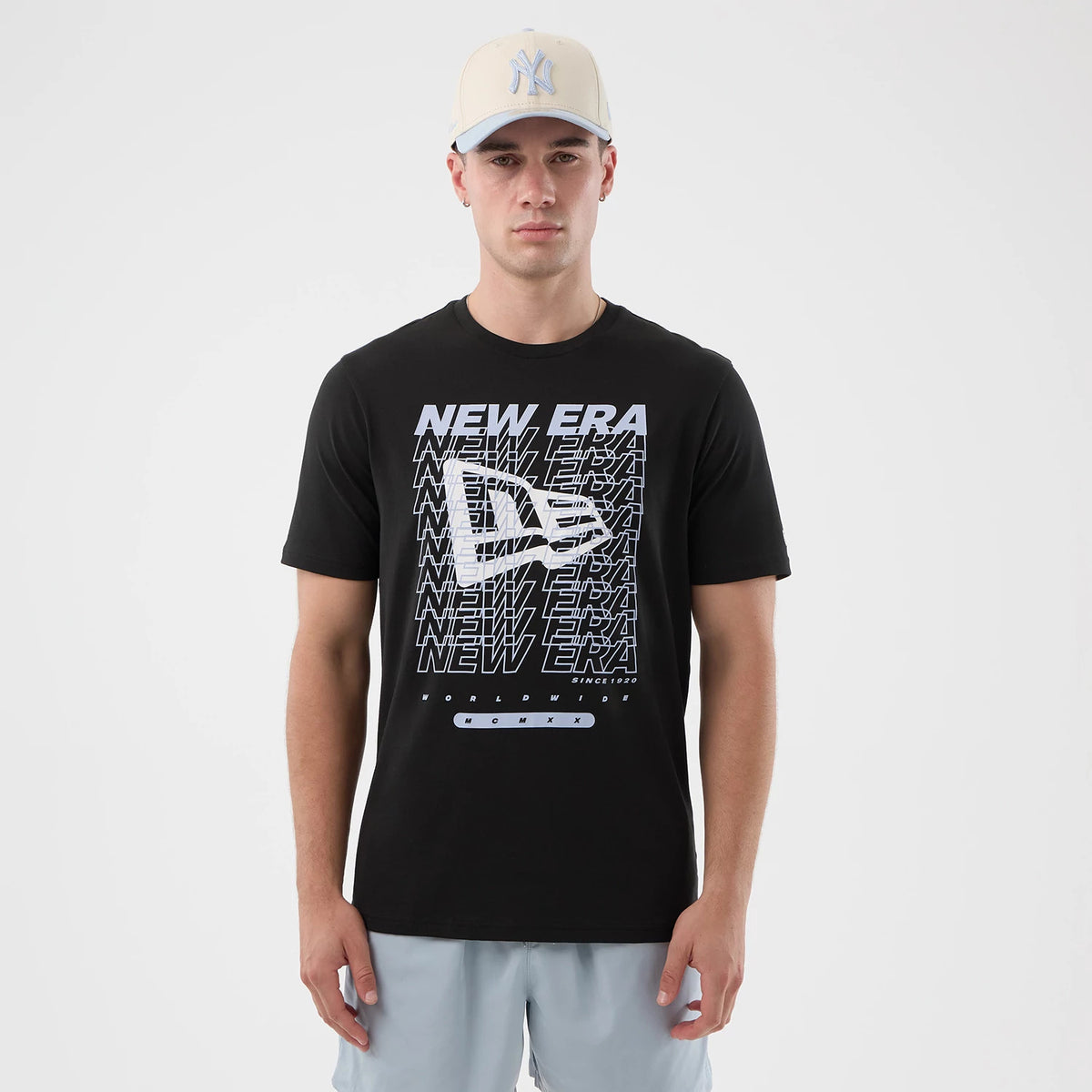 New Era Logo Graphic Black T-Shirt, front view showcasing a large white printed New Era logo pattern across the chest, with repeated lines of NEW ERA text in a grid formation, worn by a model dressed in grey shorts and a light cap.