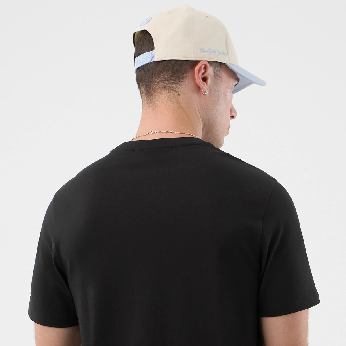 New Era Logo Graphic Black T-Shirt, back view on a model wearing a beige cap with a light blue visor, highlighting the simple black fabric of the T-shirt and showcasing a relaxed fit around the shoulders and sleeves.