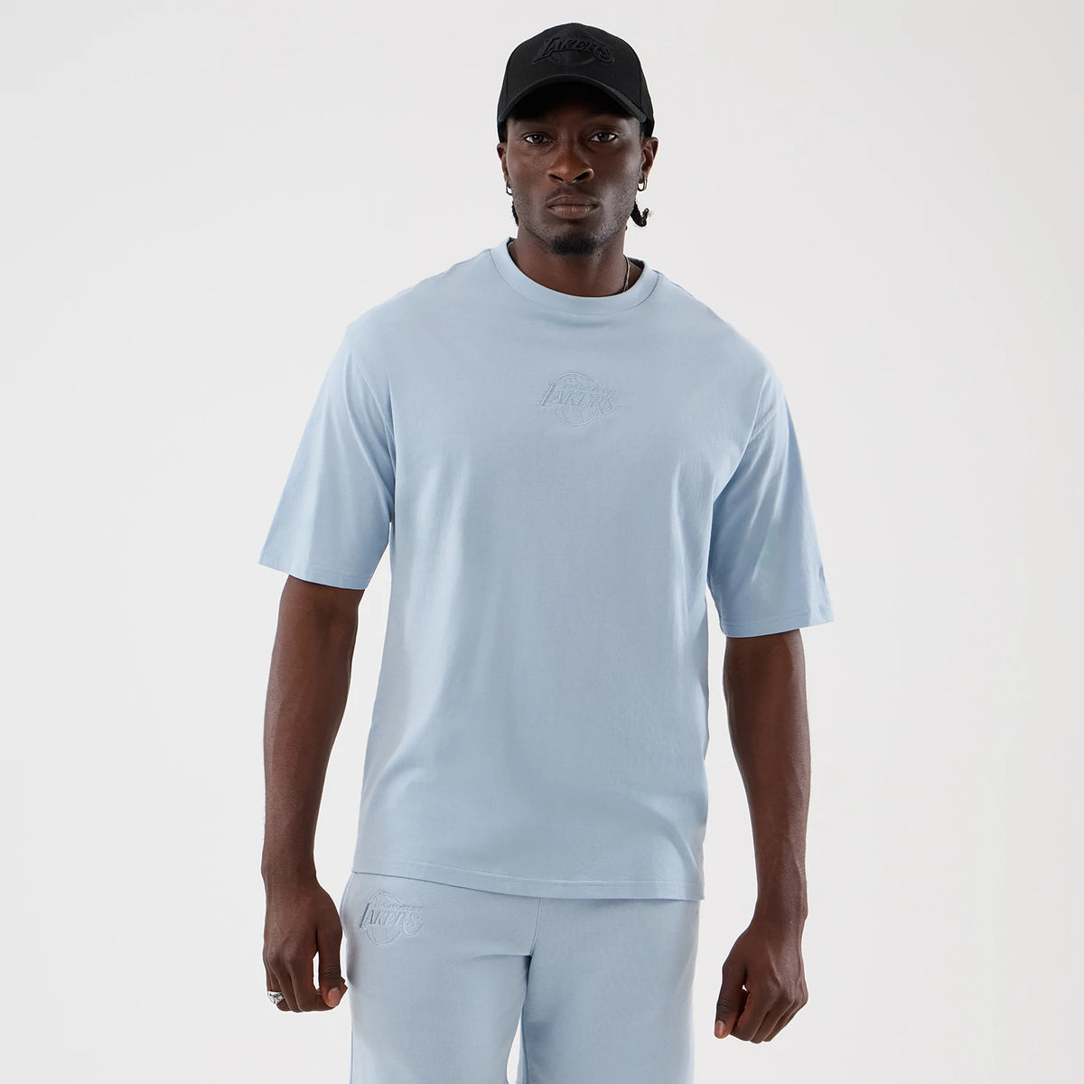 New Era LA Lakers NBA League Essential Pastel Blue Oversized T-Shirt, front view on-model featuring an embroidered Lakers logo centred on the chest, short sleeves and a relaxed fit, paired with matching shorts showing a similar logo.
