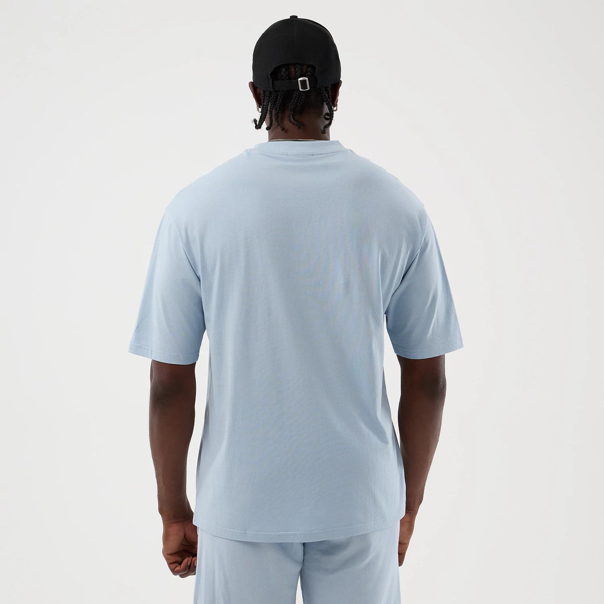 New Era LA Lakers NBA League Essential Pastel Blue Oversized T-Shirt, back view on a model wearing a black cap, displaying the plain pastel blue fabric and loose fit style that extends to the short sleeves for an oversized look.