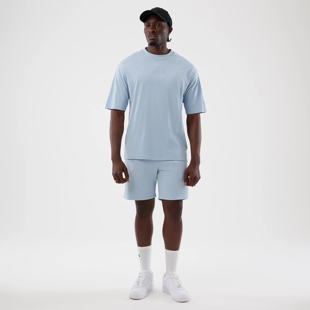 New Era LA Lakers NBA League Essential Pastel Blue Oversized T-Shirt, front view on a model featuring a embroidered Lakers logo at centre chest and relaxed fit sleeves, paired with matching pastel blue shorts and white trainers.