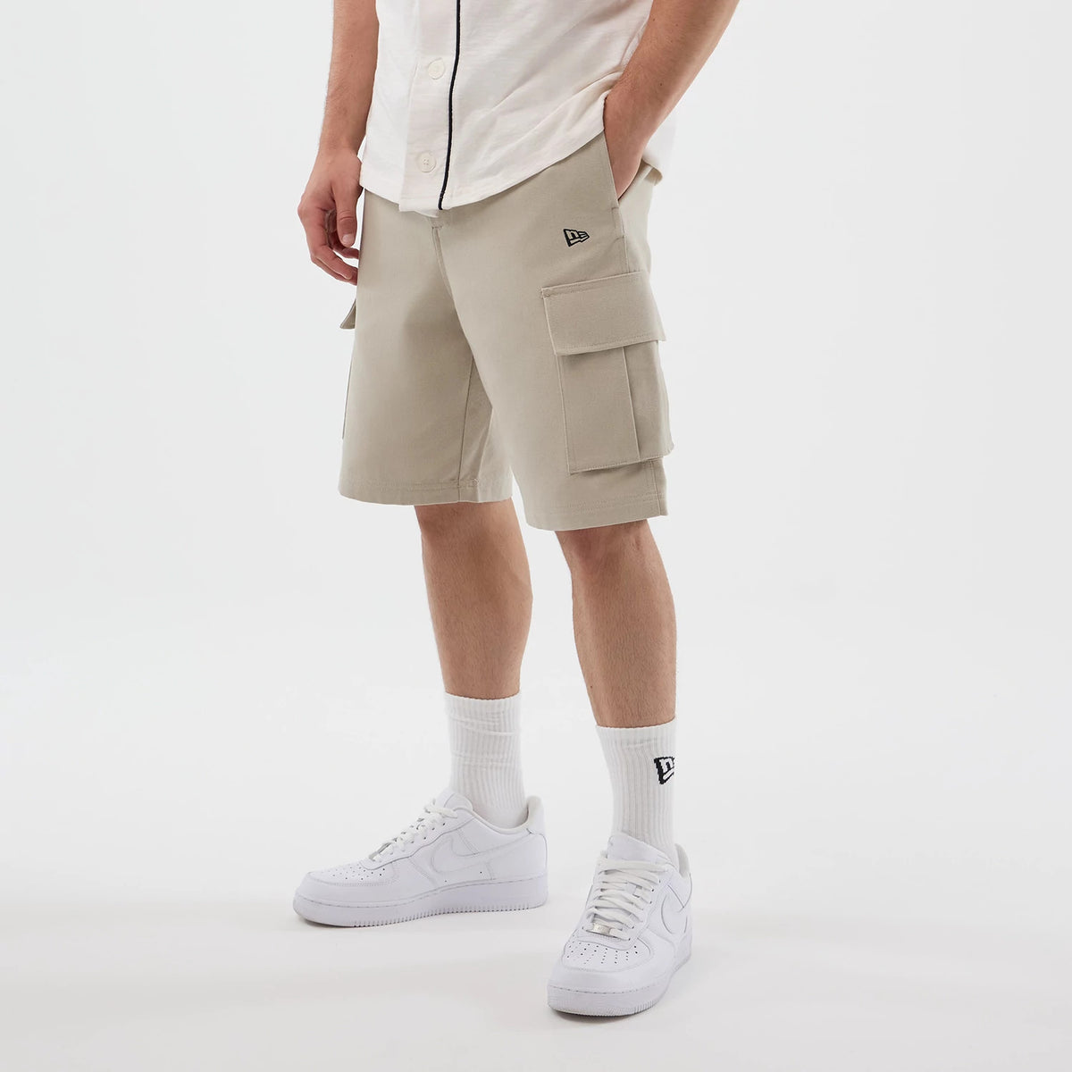 New Era Light Beige Cargo Shorts, front view on model showing the light beige colour with two large cargo pockets on each side, an embroidered New Era logo on the left pocket and paired with white socks and trainers.