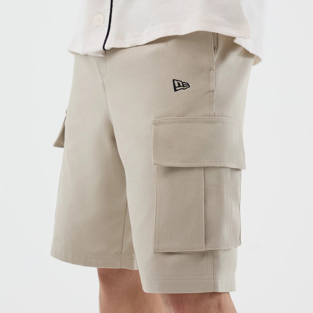 New Era Light Beige Cargo Shorts, side view showing the light beige shorts with two prominent cargo pockets on each leg, an embroidered New Era logo near the left pocket, and styled with a white shirt at the top.