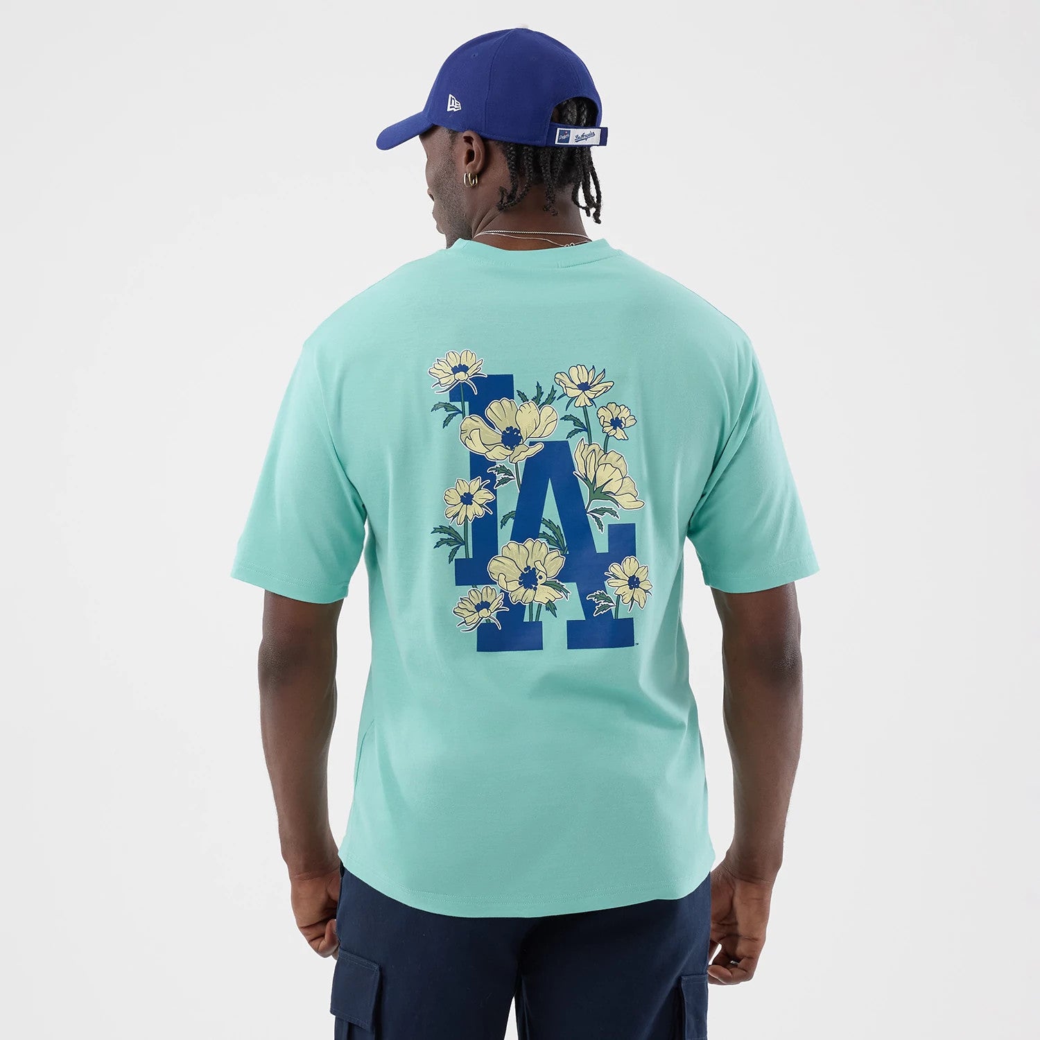 New Era LA Dodgers MLB Floral Pastel Green Oversized T-Shirt, back view on a model with blue shorts and cap, showing a large blue LA logo on the back adorned with yellow flowers and green leaves across the letters on the pastel green fabric.