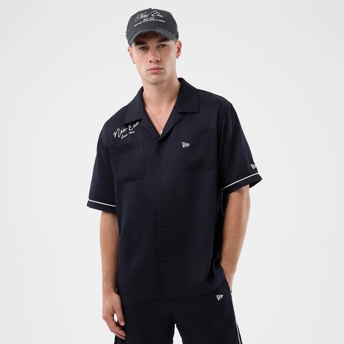 New Era Graphic Revere Navy Short Sleeve Shirt, front view on a model with matching navy shorts and cap, featuring white New Era script and the year '1920' embroidered on the left chest, and the New Era flag logo on the right chest and sleeve.