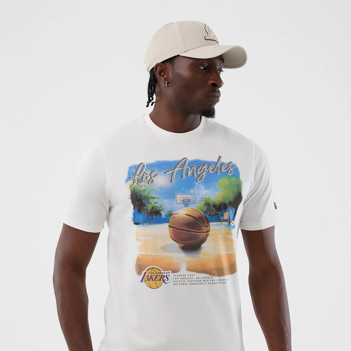 New Era LA Lakers NBA Beach Graphic White T-Shirt, front view on a model also wearing a beige cap, featuring a large printed graphic of a basketball on a beach court with palm trees and Los Angeles lettering across the chest.