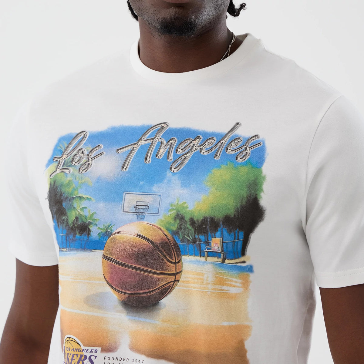 New Era LA Lakers NBA Beach Graphic White T-Shirt, front view on a model showing a colourful graphic with a basketball court, palm trees, and a large basketball, with raised script Los Angeles lettering across the chest.