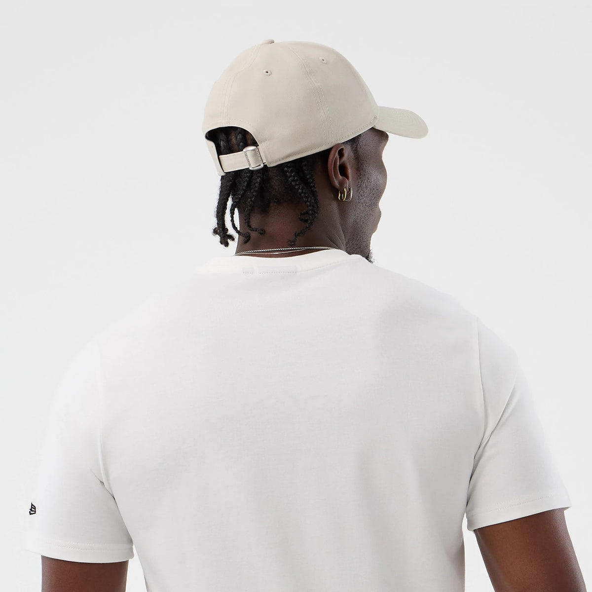 New Era LA Lakers NBA Beach Graphic White T-Shirt, back view on a model showing a plain white design, short sleeves with a small embroidered New Era logo on the left sleeve, paired with a beige cap featuring an adjustable strap and eyelets.