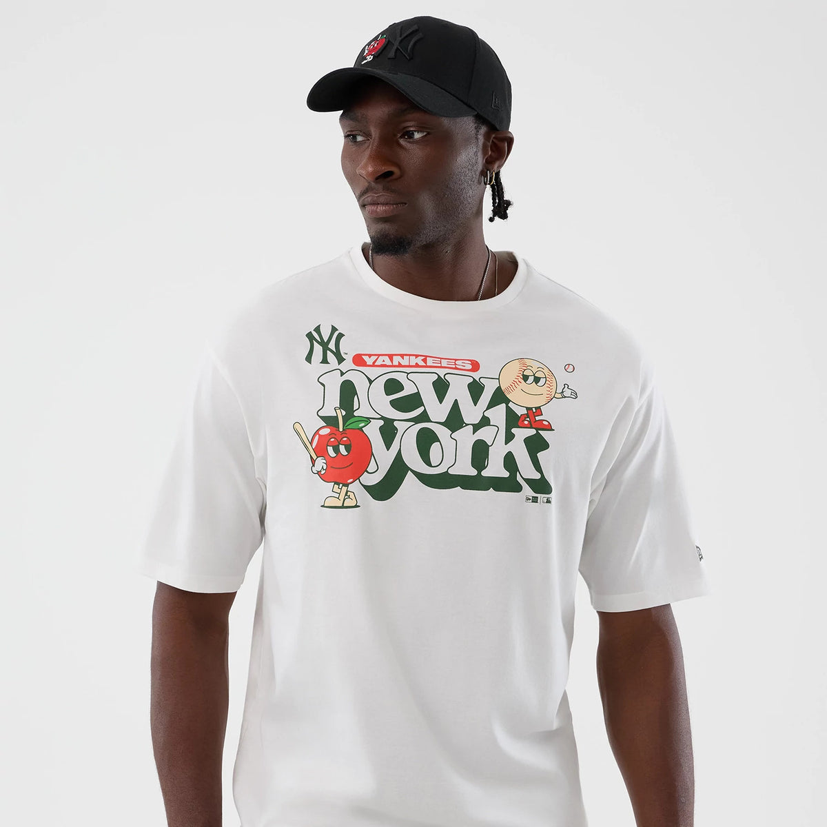 New Era New York Yankees MLB Icon Graphic White T-Shirt, front view on model with matching black cap, displaying large green new york text, cartoon apple and baseball characters, and NY Yankees logo printed above the graphics on a white fabric.