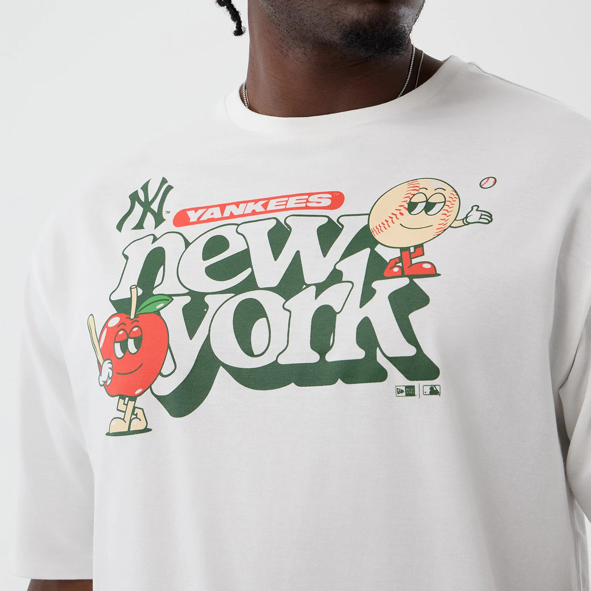 New Era New York Yankees MLB Icon Graphic White T-Shirt, front view displaying a graphic with new york in large green lettering, featuring cartoon characters of an apple and baseball, and the Yankees logo printed near the upper left shoulder.