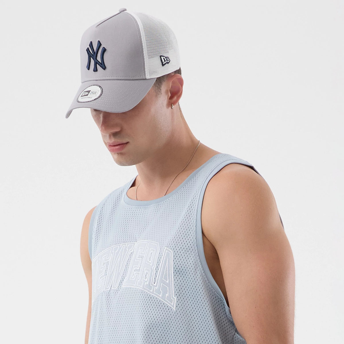 New Era Mesh Pastel Blue Oversized Tank Top, on-model view paired with a grey cap, showing a large white New Era logo on the chest of the tank top made of lightweight mesh, with sleeveless design and round neckline.