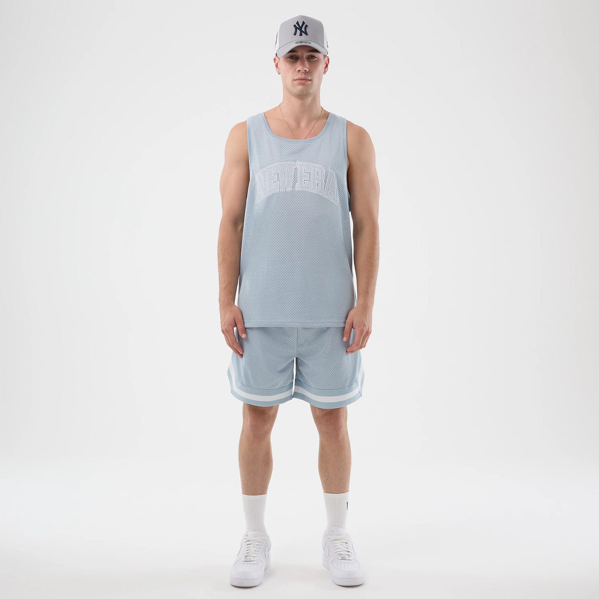 New Era Mesh Pastel Blue Oversized Tank Top, front view on a model wearing a matching cap, showcasing the white embroidered New Era lettering across the chest on a light blue mesh fabric, paired with coordinating pastel blue shorts.