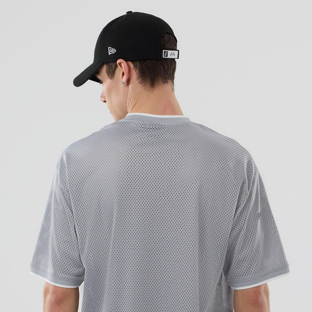 New Era San Antonio Spurs NBA Mesh Grey Oversized T-Shirt, back view on a model wearing a black cap with an embroidered logo, showing the grey mesh material, white neckline and sleeve trim, and seamless design across the shoulders.