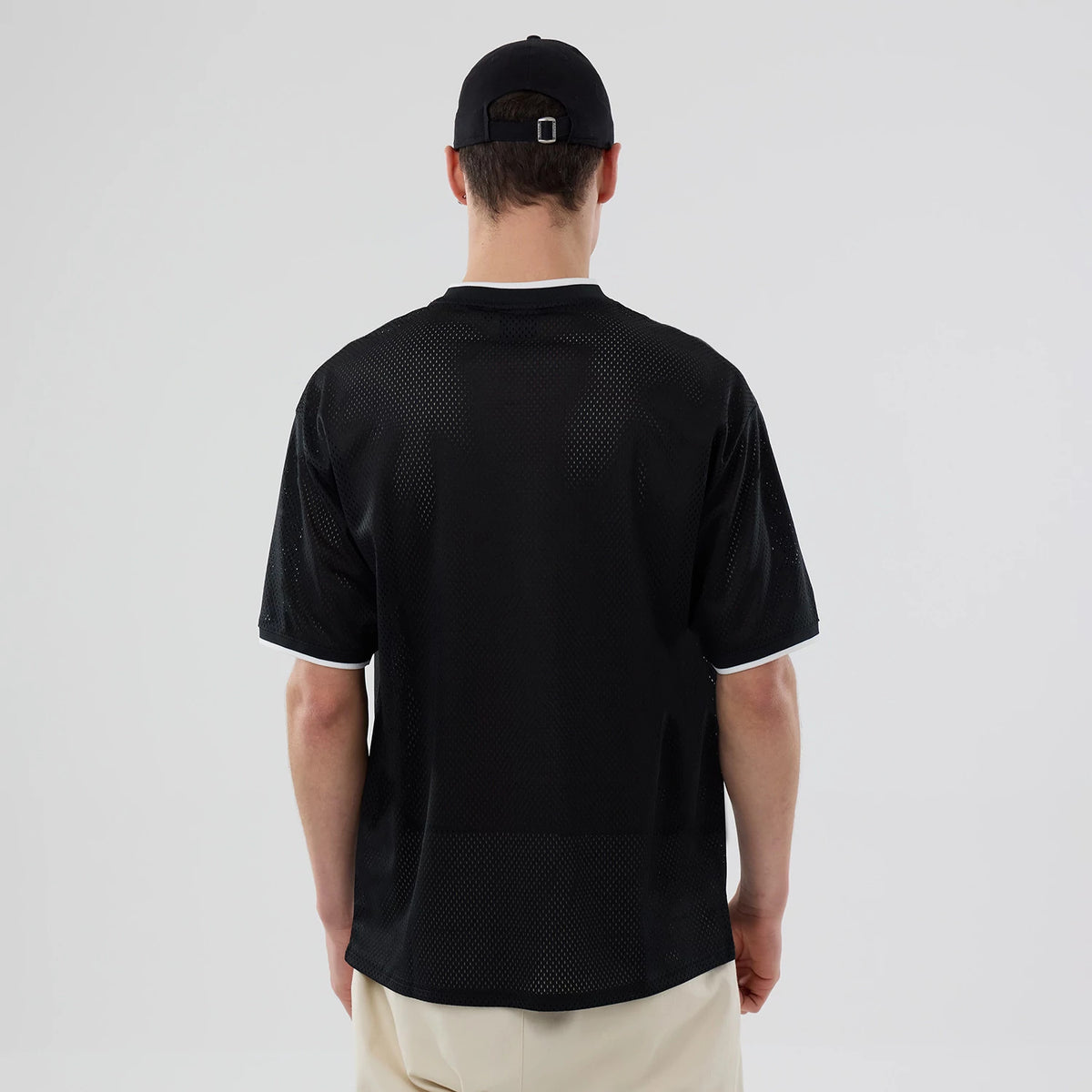 New Era Brooklyn Nets NBA Mesh Black Oversized T-Shirt, back view on a model wearing a black cap and beige trousers, showcasing the plain black mesh design with white trim on the cuffs and collar, revealing a casual yet sporty look.
