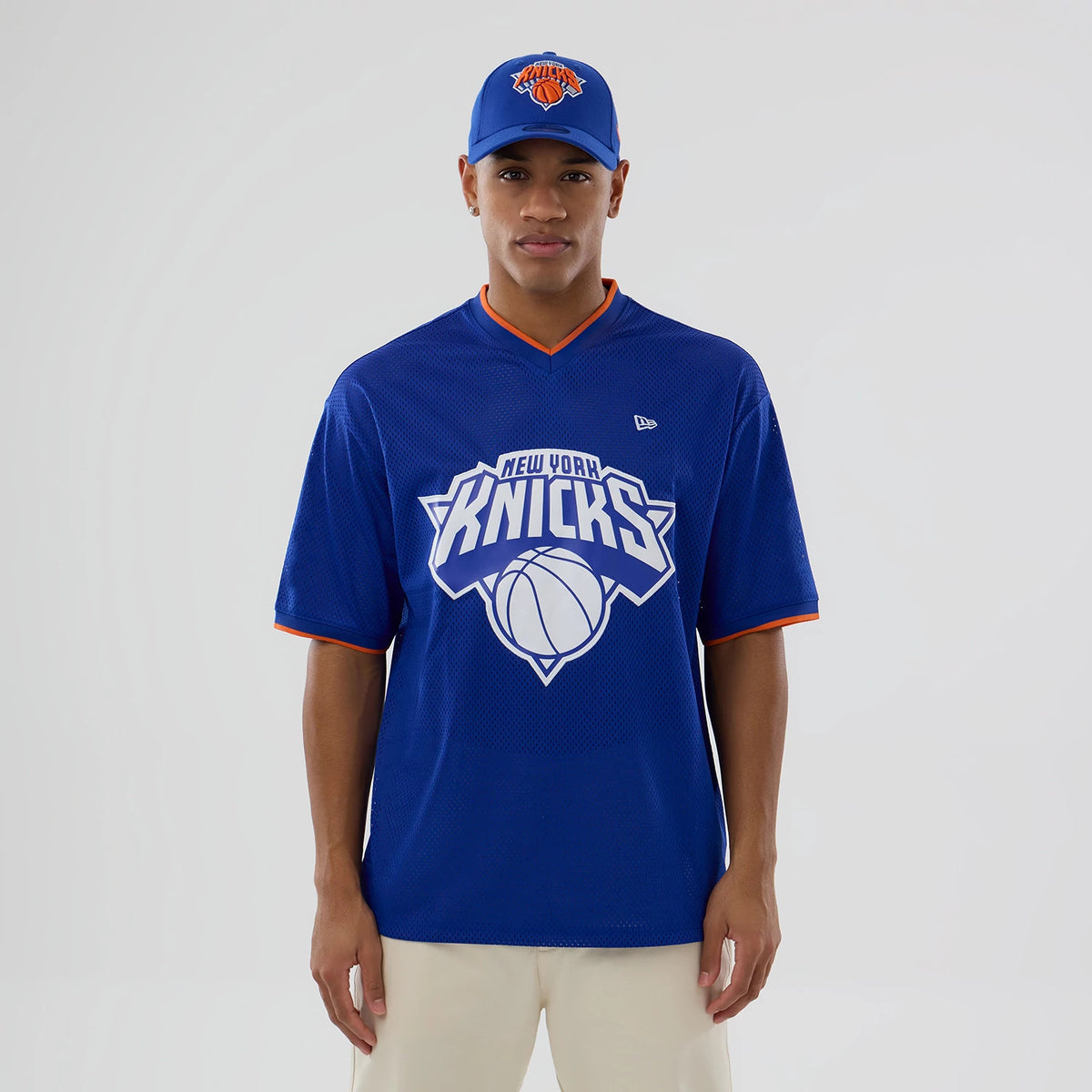New Era New York Knicks NBA Mesh Blue Oversized T-Shirt, front view on a model wearing a matching cap, displaying a large printed Knicks basketball logo across the chest, a small embroidered New Era logo on the upper right chest.