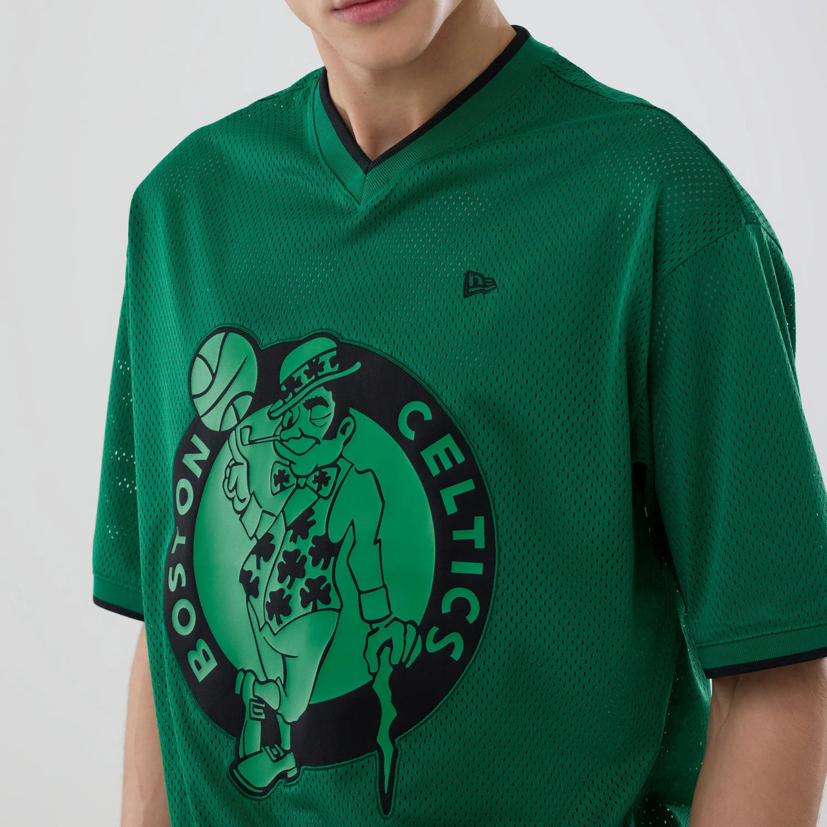 New Era Boston Celtics NBA Mesh Green Oversized T-Shirt, front view showing a large graphic of the Celtics logo centred on the chest, a small black embroidered New Era logo on the upper left, and a black-trimmed V-neckline on the green mesh fabric.