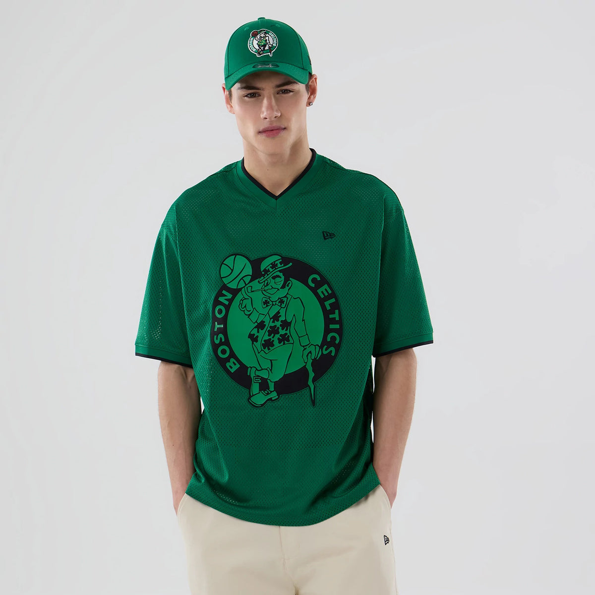 New Era Boston Celtics NBA Mesh Green Oversized T-Shirt, front view on-model showing a large Celtics graphic centred on the green mesh fabric, New Era logo embroidered on the left chest, model with matching green cap.