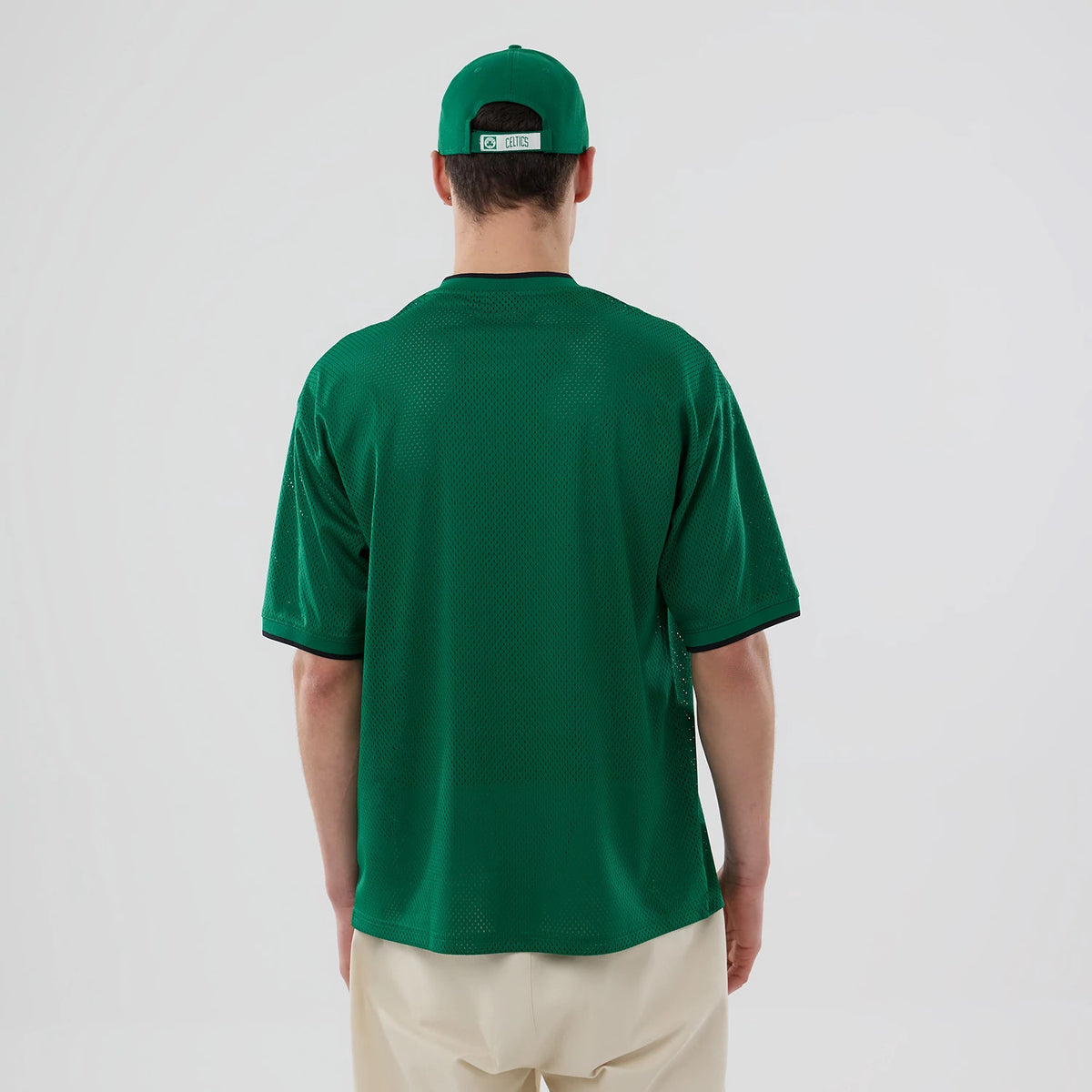 New Era Boston Celtics NBA Mesh Green Oversized T-Shirt, back view on a model paired with a matching green cap, featuring a plain green mesh texture with black edging around the collar and sleeves, giving a clean and sporty appearance.