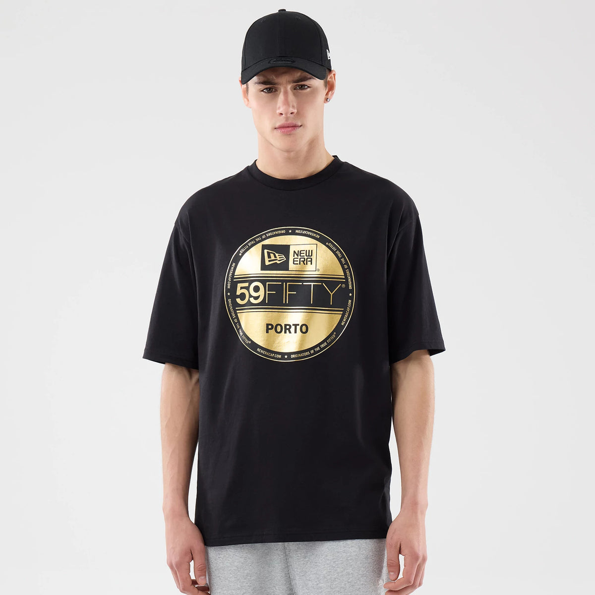 New Era Sticker Porto Black Oversized T-Shirt, front view on a model styled with a black cap, featuring a large gold circular graphic on the chest with 59FIFTY lettering, Porto text below, and a printed New Era logo at the top.
