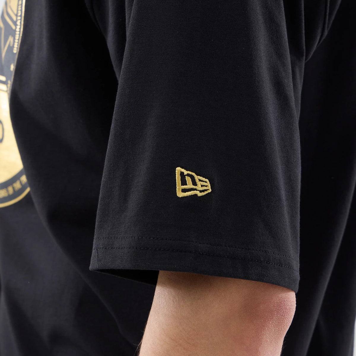 New Era Sticker Porto Black Oversized T-Shirt, on-model view focusing on the sleeve showing a small embroidered gold New Era logo near the end of the sleeve and part of a large circular graphic in gold and cream on the black fabric.