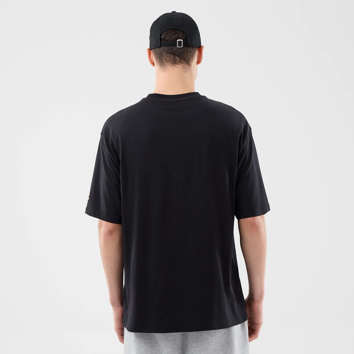 New Era Sticker Dubai Black Oversized T-Shirt, back view on a model showing the plain oversized black back with short sleeves, worn with a black cap featuring an adjustable strap and buckle, styled with light grey shorts.
