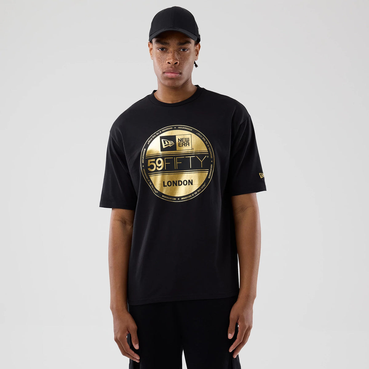 New Era Sticker London Black Oversized T-Shirt, front view displaying a large gold printed circular graphic of the New Era 59FIFTY logo and London text on the chest, with a small gold embroidered New Era logo on the left sleeve, worn by a model.