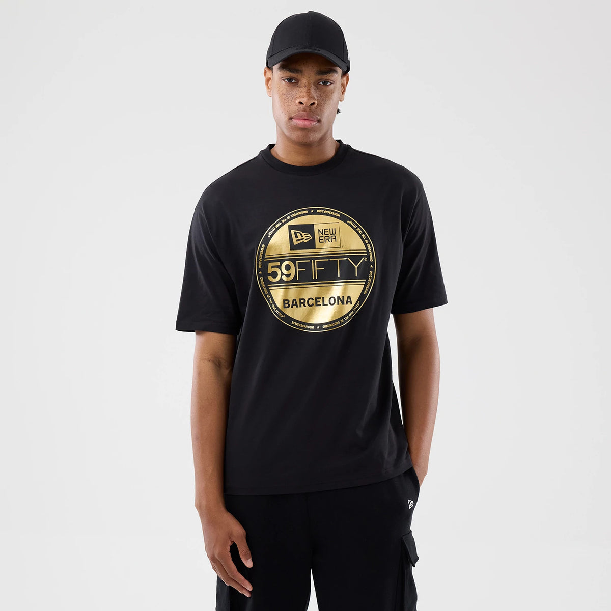 New Era Sticker Barcelona Black Oversized T-Shirt, front view on a model with a matching black cap, featuring a large gold New Era 59FIFTY logo print at the centre of the chest and a relaxed fit with short sleeves.