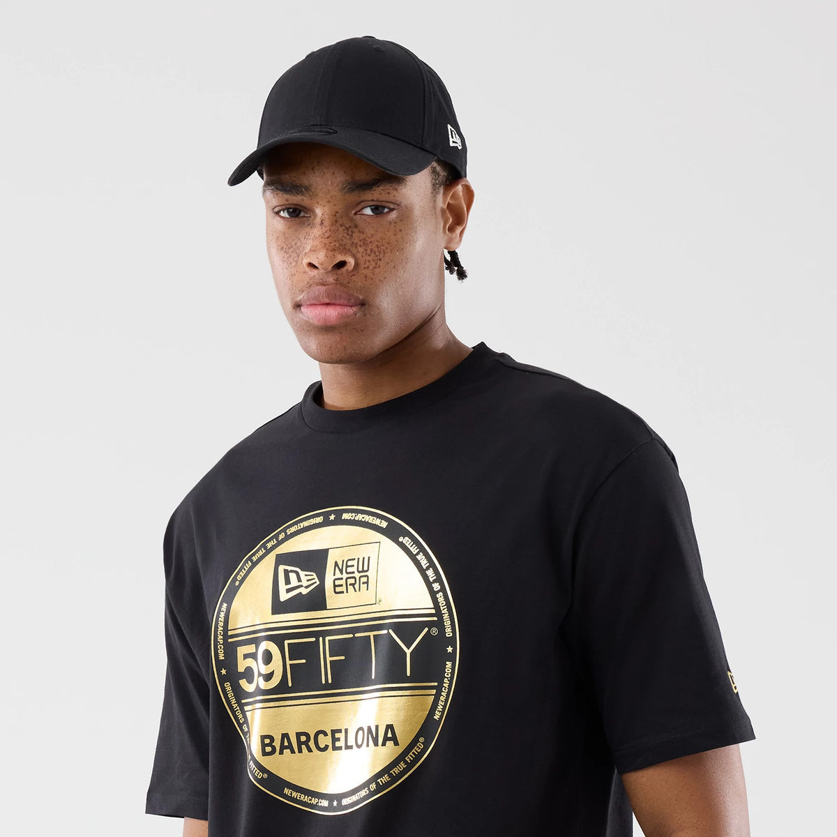 New Era Sticker Barcelona Black Oversized T-Shirt, front view on a model with a plain black cap, featuring a large circular gold print with New Era and 59FIFTY branding across the chest and a New Era logo on the left sleeve.