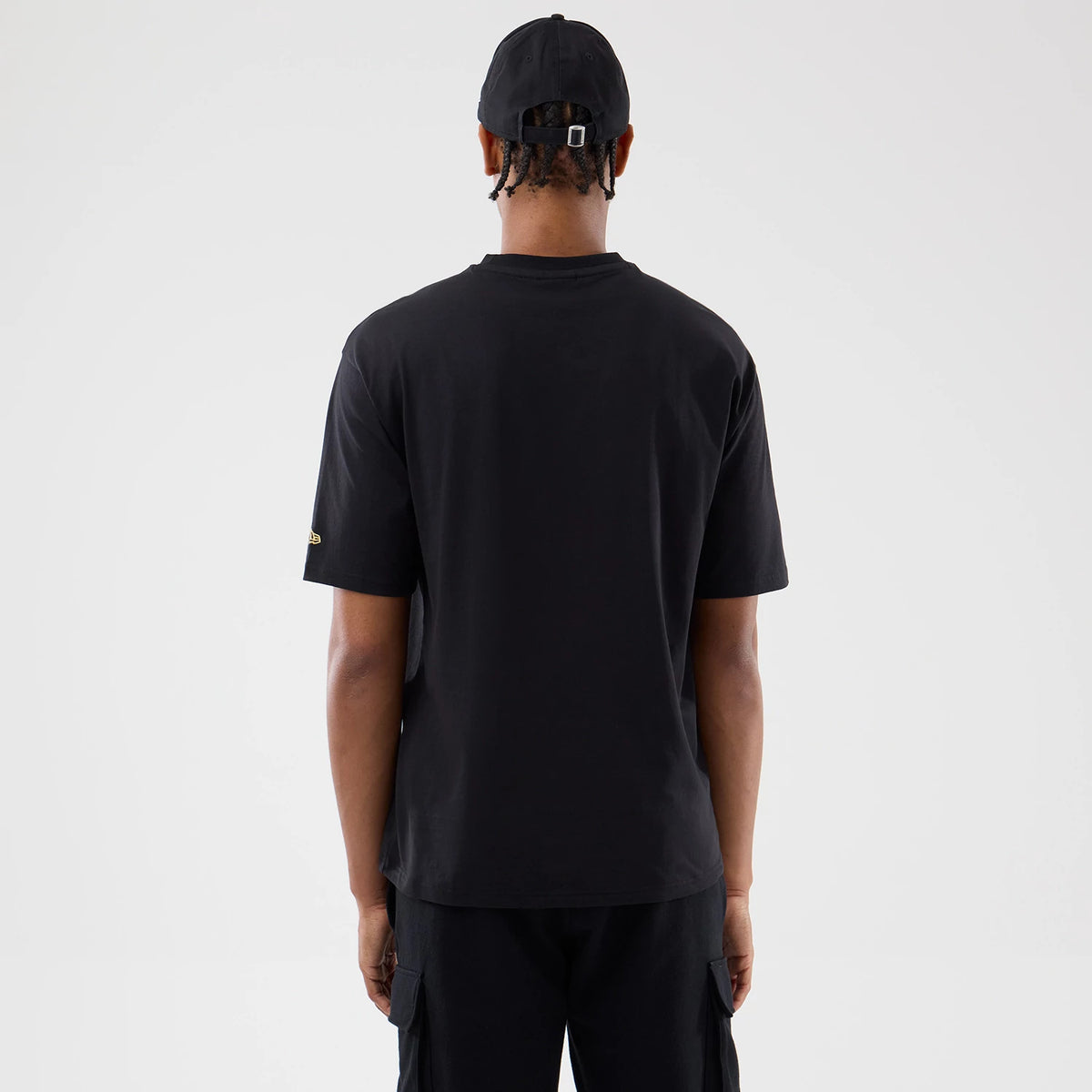 New Era Sticker Barcelona Black Oversized T-Shirt, back view on a model wearing matching black trousers and a cap, showing a plain black back with a small gold-embroidered New Era logo on the left sleeve.