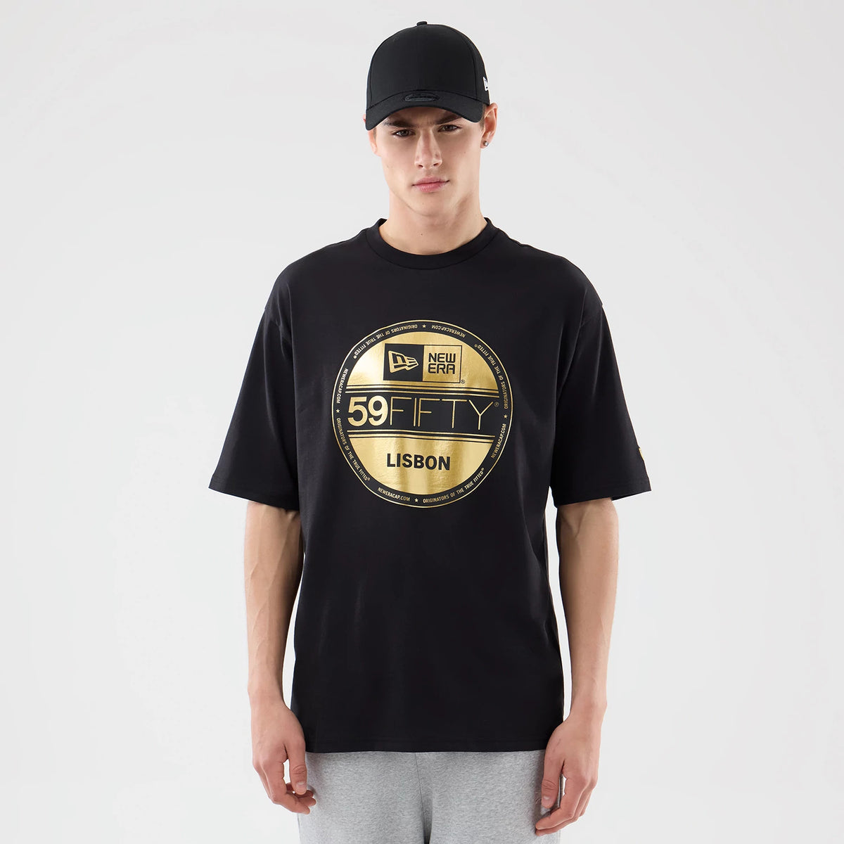 New Era Sticker Lisbon Black Oversized T-Shirt, front view on a model wearing a black cap, featuring a large gold printed New Era 59FIFTY logo and Lisbon lettering across the chest, with short sleeves and a relaxed fit.